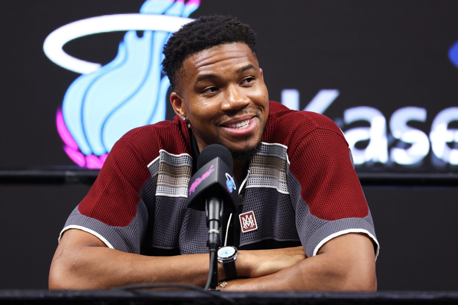 Giannis Antetokounmpo speaks into microphones with a Miami Heat logo behind him.
